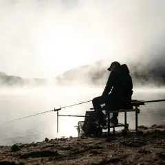 Wędkarz siedzi nad mglistym jeziorem o wschodzie słońca, łowiąc ryby.