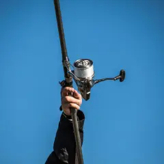 Dłoń trzyma wędkę z nawiniętą plecionką na kołowrotku, na tle błękitnego nieba.