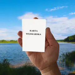 Dłoń trzyma kartkę z napisem "KARTA WĘDKARSKA". Czy egzamin na kartę wędkarską online to prawda czy mit? Piękny krajobraz nad jeziorem.