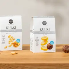 Dwa opakowania kulek proteinowych: krem orzechowy i banan, obok nich owoce i orzeszki.