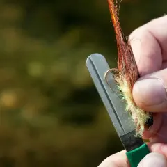 Ręka ostrzy haczyk muchowy na kamieniu, pokazując jak ostrzyć tępe haczyki nad wodą bez specjalnej osełki.