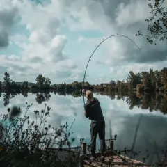 Wędkarz na pomoście łowi ryby na spokojnym jeziorze, z drzewami w tle.