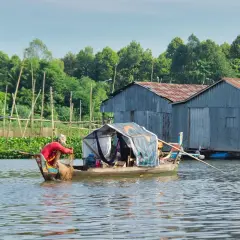 Rybak w łodzi z siecią na tle domów na wodzie i zieleni.