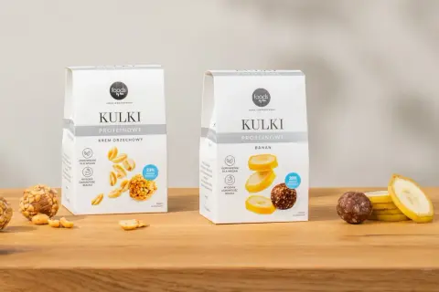 Dwa opakowania kulek proteinowych: krem orzechowy i banan, obok nich owoce i orzeszki.
