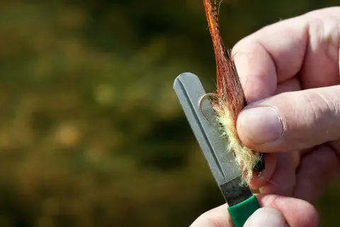 Ręka ostrzy haczyk muchowy na kamieniu, pokazując jak ostrzyć tępe haczyki nad wodą bez specjalnej osełki.