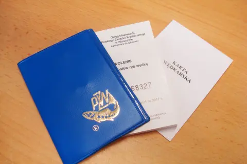 Niebieska okładka karty wędkarskiej z logo ryby i złotym napisem PZW, obok dokumenty.