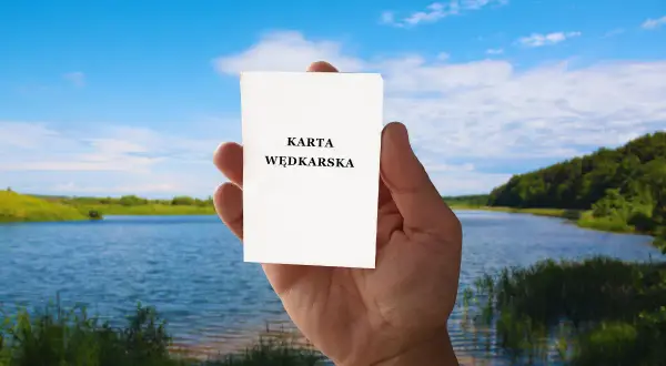 Dłoń trzyma kartkę z napisem "KARTA WĘDKARSKA". Czy egzamin na kartę wędkarską online to prawda czy mit? Piękny krajobraz nad jeziorem.