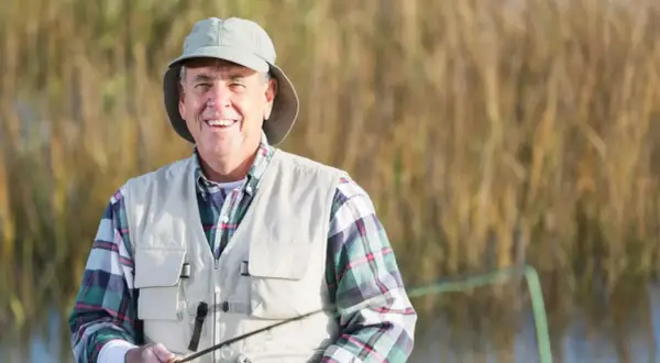 Uśmiechnięty wędkarz w kapeluszu i kamizelce, cieszący się wędkowaniem. Posiada kartę wędkarską dla osoby niepełnosprawnej, co zapewnia ulgi i zwolnienia z opłat.
