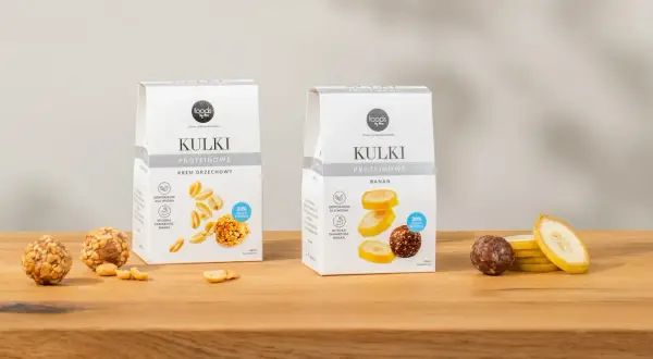Dwa opakowania kulek proteinowych: krem orzechowy i banan, obok nich owoce i orzeszki.
