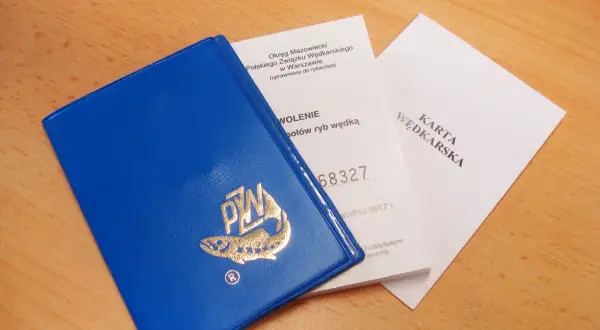 Niebieska okładka karty wędkarskiej z logo ryby i złotym napisem PZW, obok dokumenty.