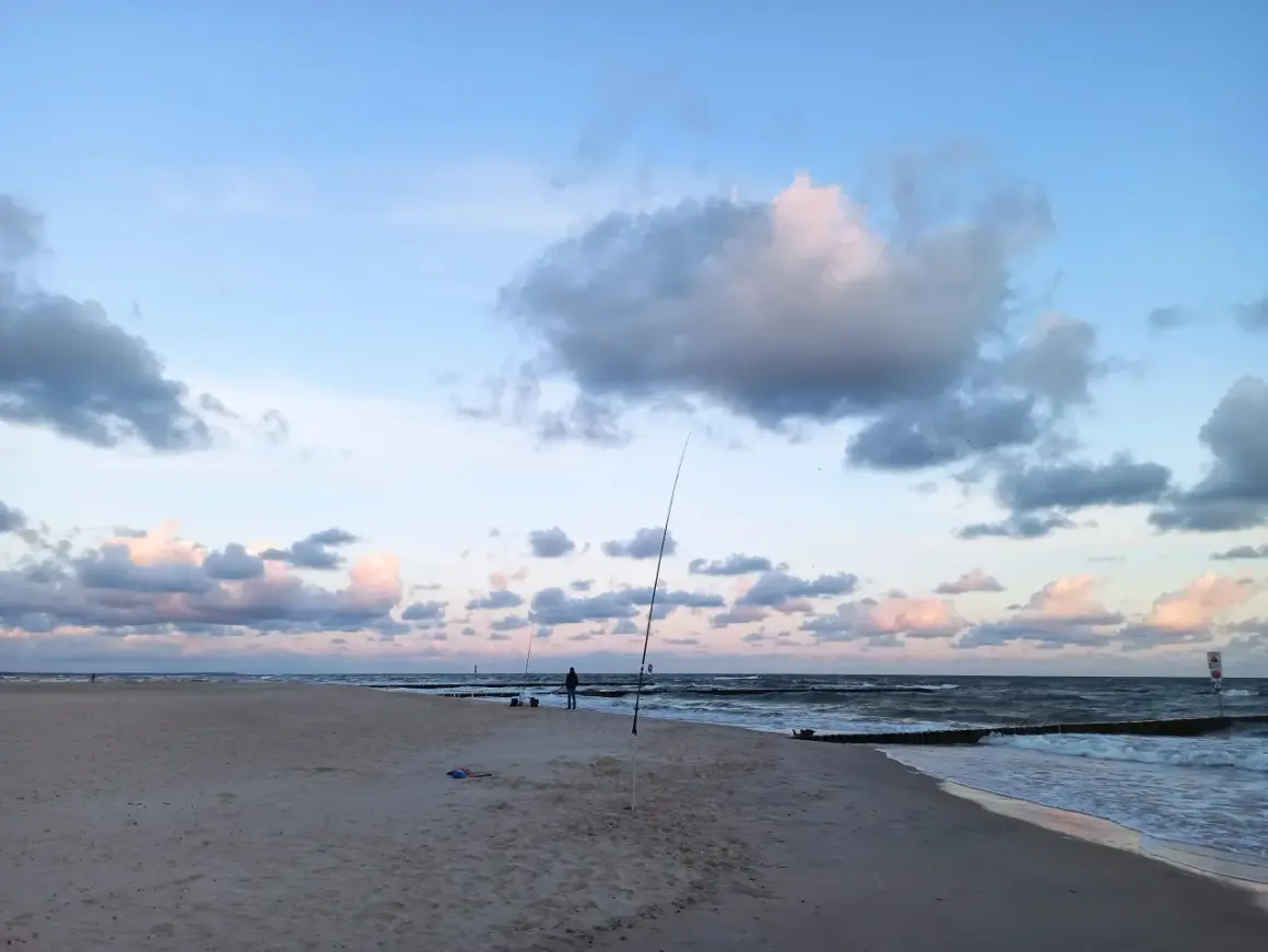 Surfcasting z plaży nad Bałtykiem. Wędkarz przygotowuje zestaw na dalekie rzuty, w tle fale i chmury.