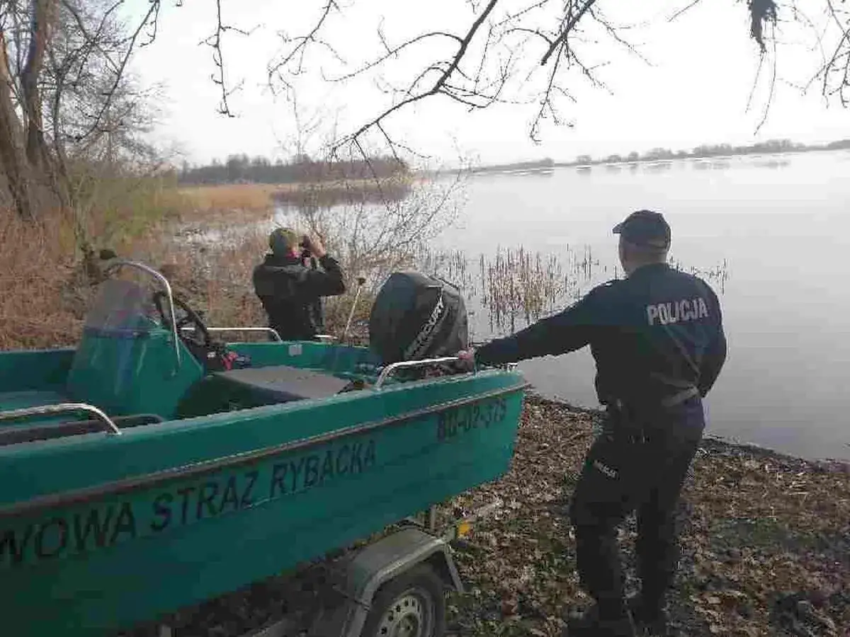 Policjant dokonuje weryfikacji autentyczności karty wędkarskiej podczas kontroli nad wodą. Obok łódź Straży Rybackiej.