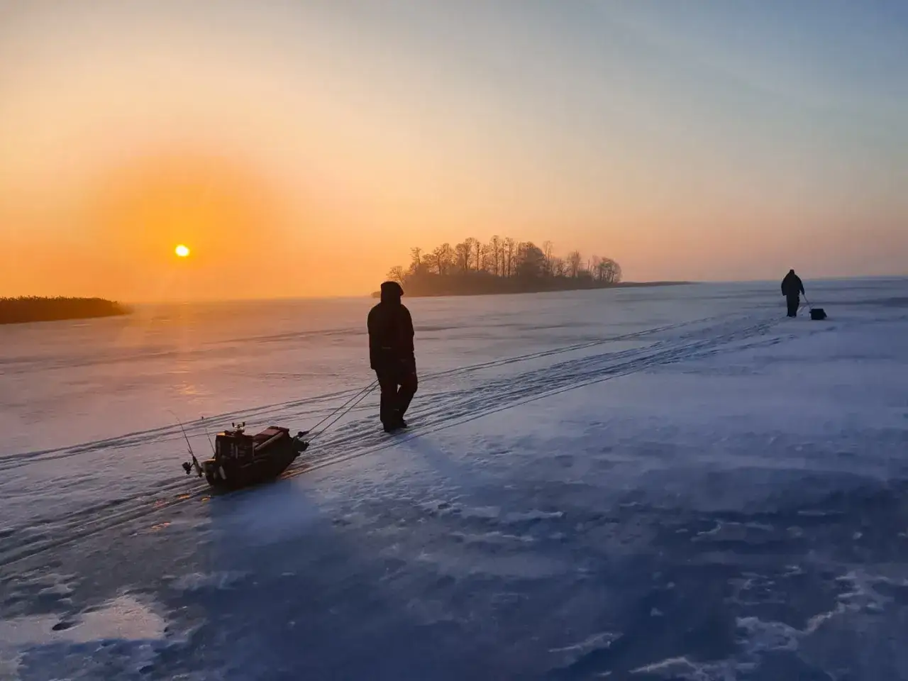 Wschód słońca nad zamarzniętym jeziorem. Wędkarz ciągnie sanie z ekwipunkiem, przygotowując się do łowienia na mormyszkę bez ochotki czy bezmotylka, co jest skuteczne na okonie.