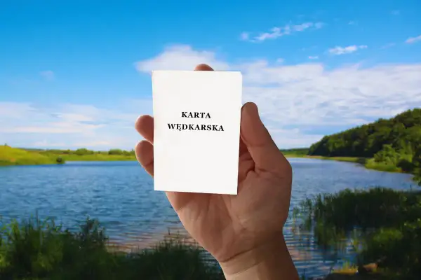 Dłoń trzyma kartkę z napisem "KARTA WĘDKARSKA". Czy egzamin na kartę wędkarską online to prawda czy mit? Piękny krajobraz nad jeziorem.