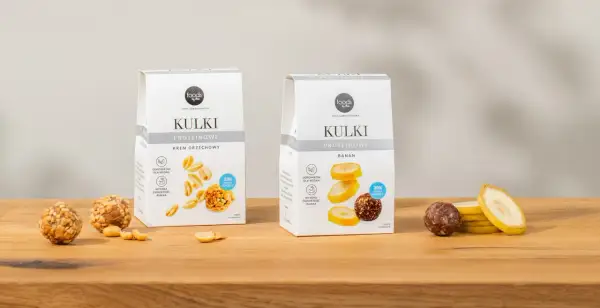 Dwa opakowania kulek proteinowych: krem orzechowy i banan, obok nich owoce i orzeszki.