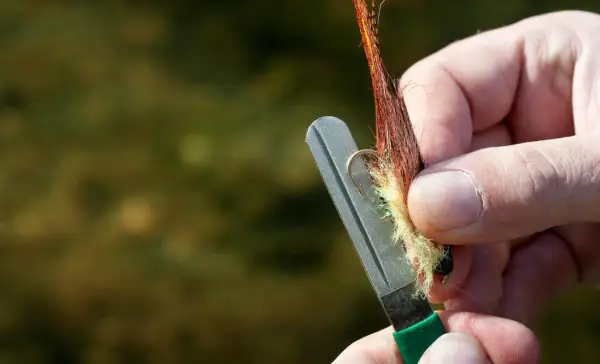 Ręka ostrzy haczyk muchowy na kamieniu, pokazując jak ostrzyć tępe haczyki nad wodą bez specjalnej osełki.