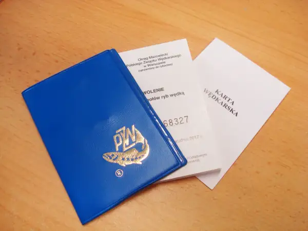Niebieska okładka karty wędkarskiej z logo ryby i złotym napisem PZW, obok dokumenty.